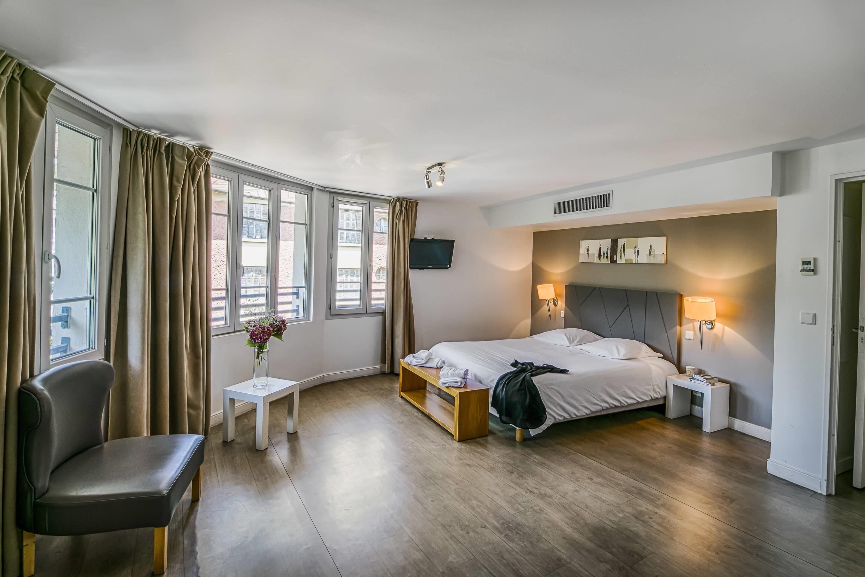 Ein Hotelzimmer mit einem Bett, Nachttischen, einem Sessel, einem kleinen Tisch mit Blumenvase und einem an der Wand montierten Fernseher.