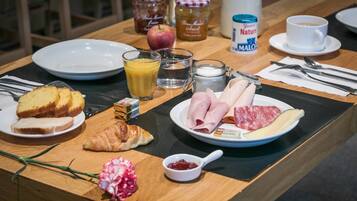 Une table de petit-déjeuner avec des assiettes remplies de nourriture, notamment des croissants, du jambon et du fromage, ainsi que des condiments, un verre de jus d’orange et une tasse de café.