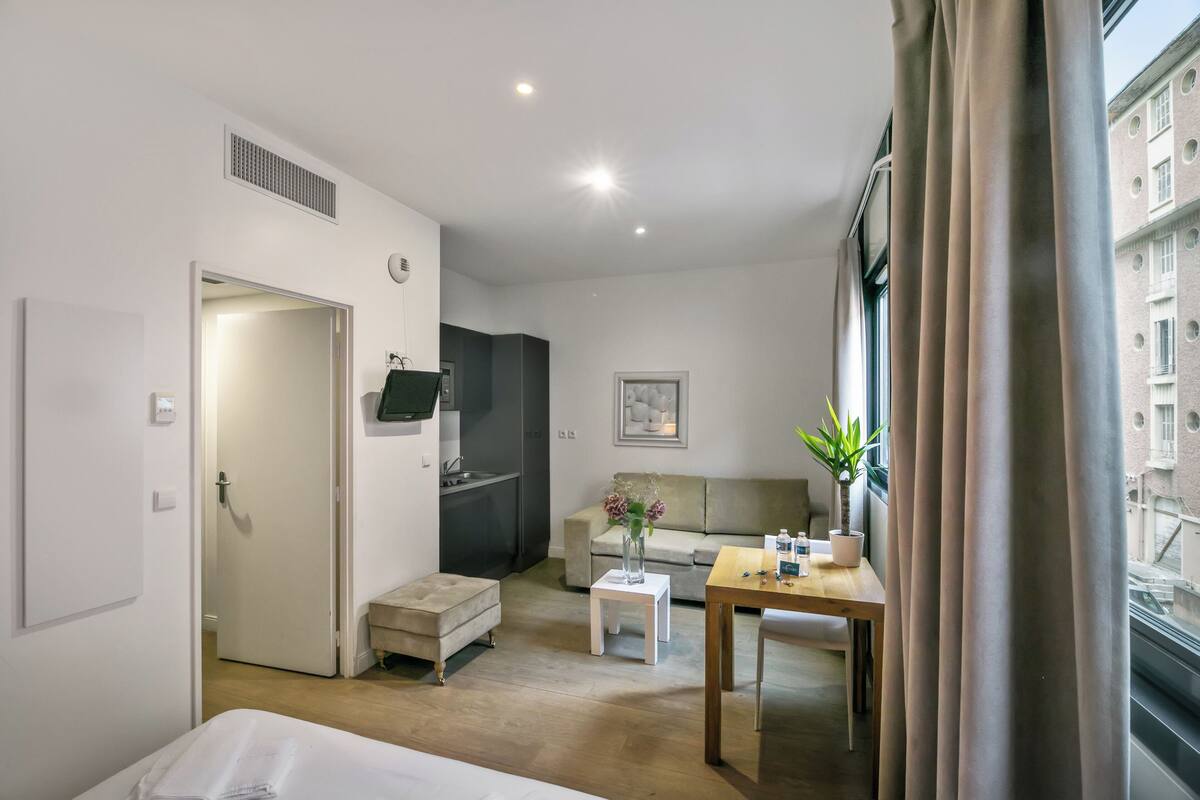 Une chambre d’hôtel avec un lit, une petite table, un canapé, une kitchenette et une fenêtre avec des rideaux.
