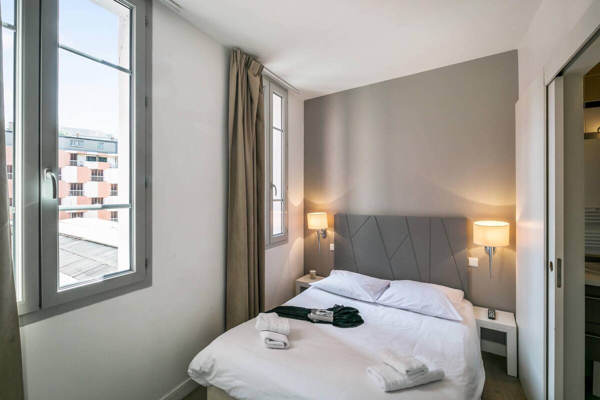 Une chambre d’hôtel avec un lit, deux lampes de chevet, une fenêtre avec des rideaux et une vue sur les bâtiments environnants.