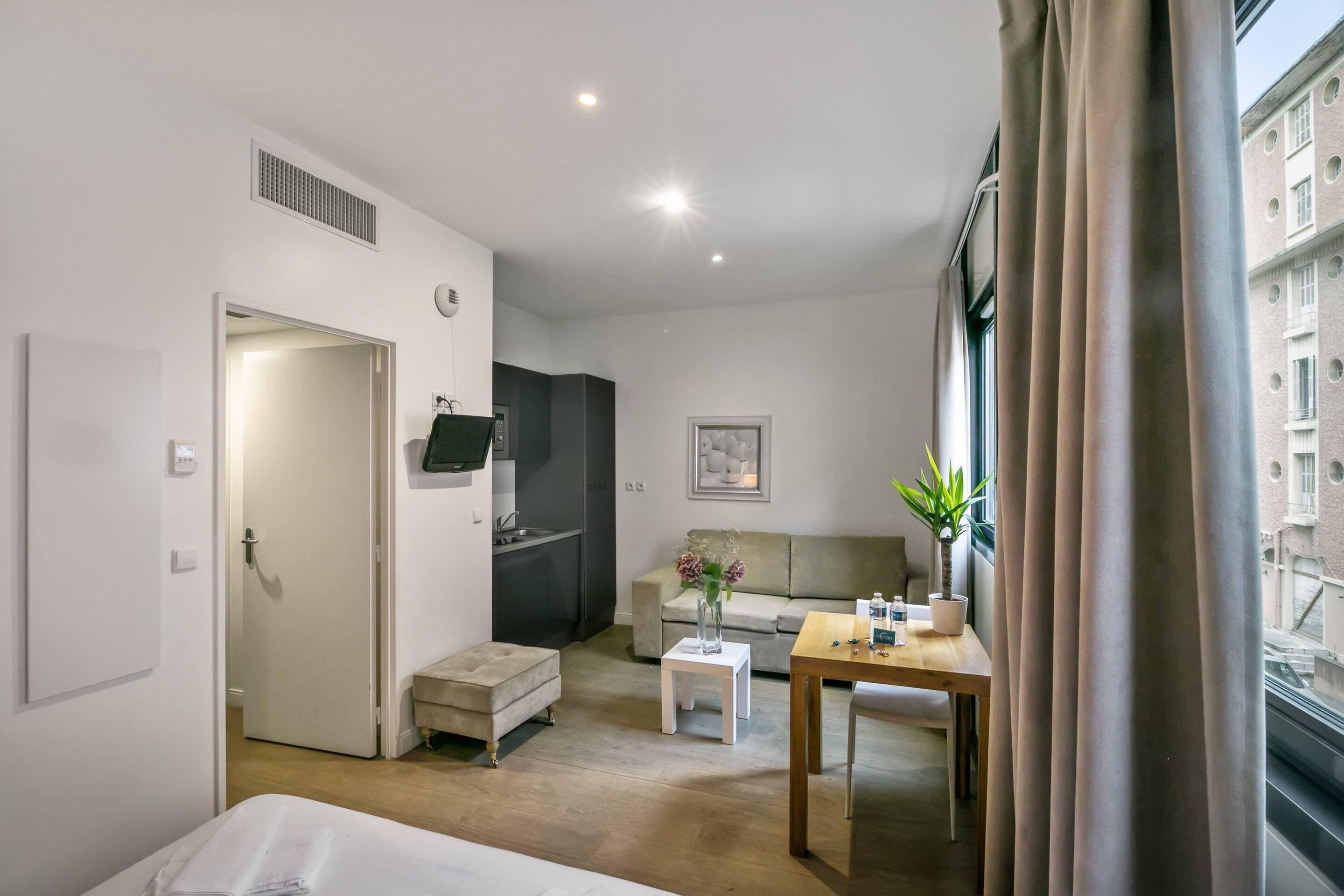 A hotel room with a bed, a small table, a sofa, a potted plant, and a window with curtains.