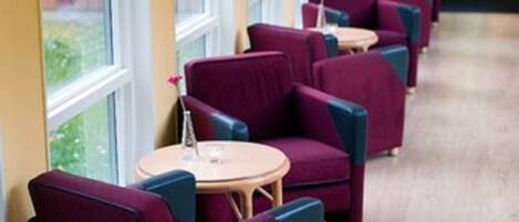 A row of purple armchairs and small round tables in a hallway with large windows and wooden flooring.