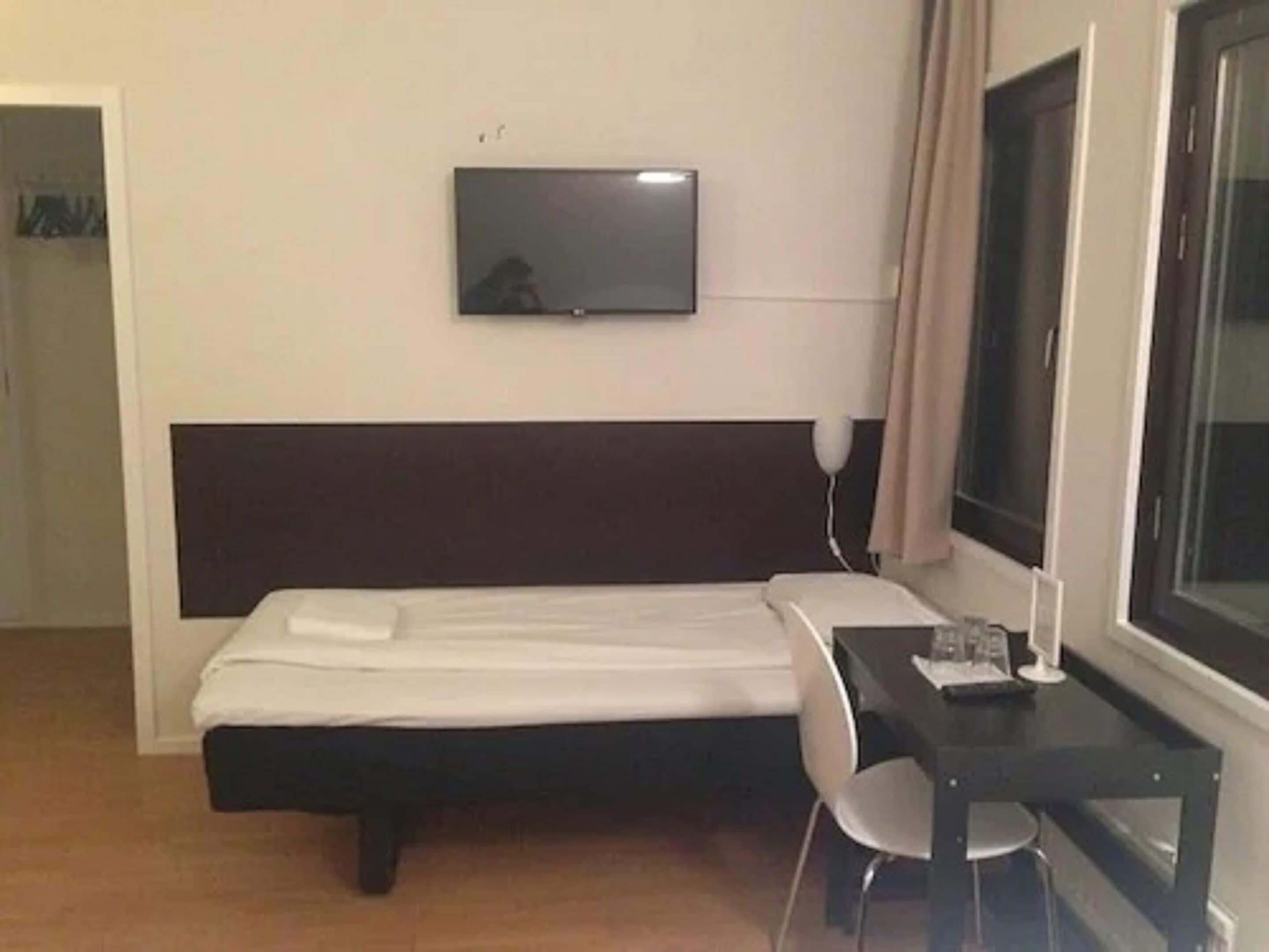 Een hotelkamer met een bed, een bureau, een stoel, een aan de muur bevestigde televisie en een raam met gordijnen.