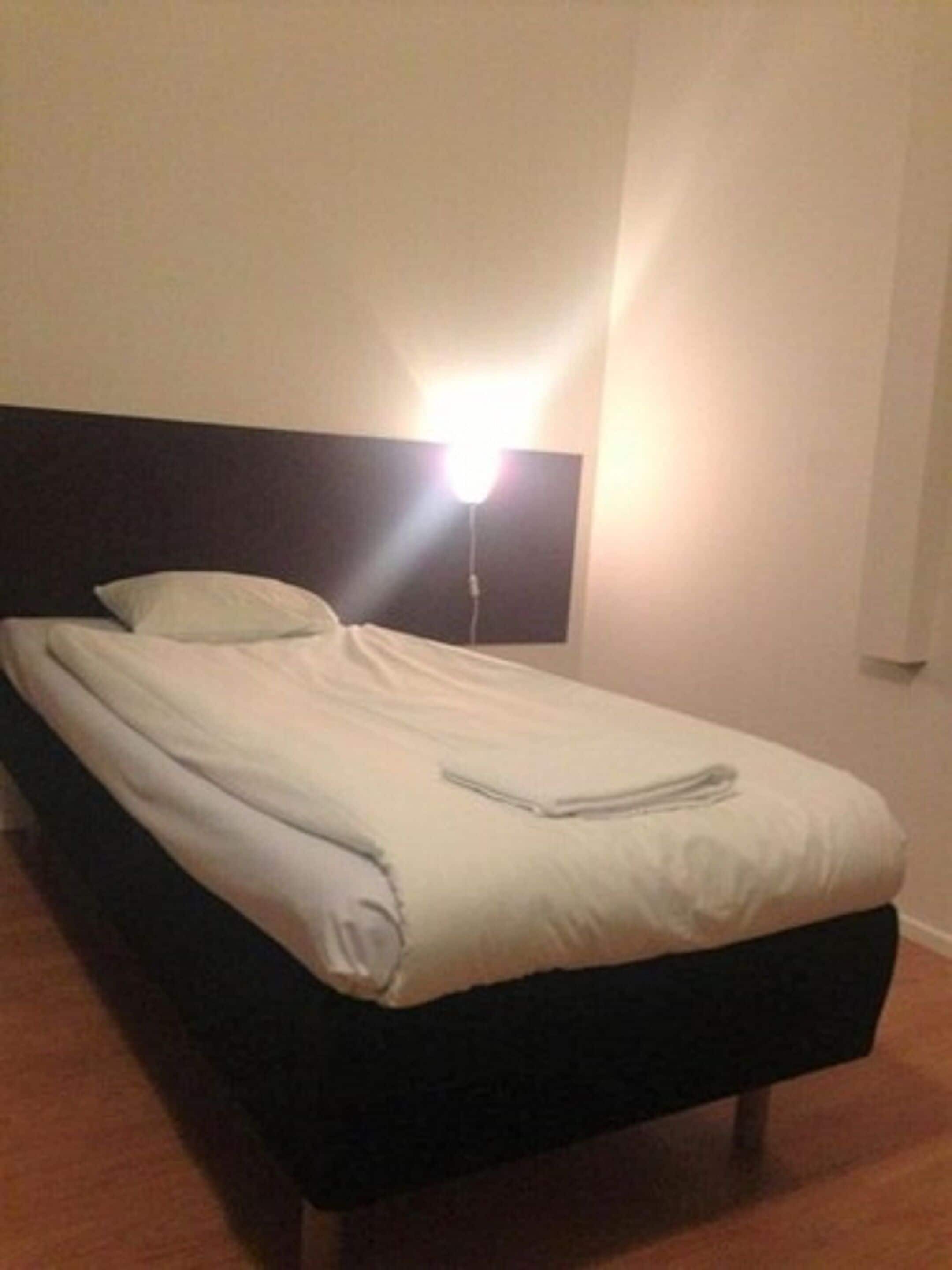 Una cama bien hecha con sábanas blancas y un cabecero oscuro en una habitación con pared blanca lisa y suelo de madera.