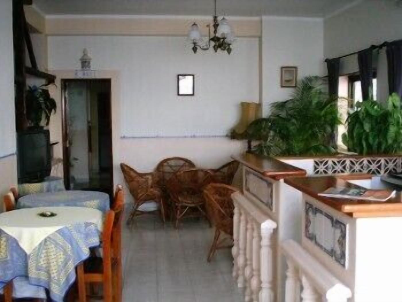 Un comedor con una mesa redonda, sillas y una araña. Hay plantas y una mesita con objetos encima.