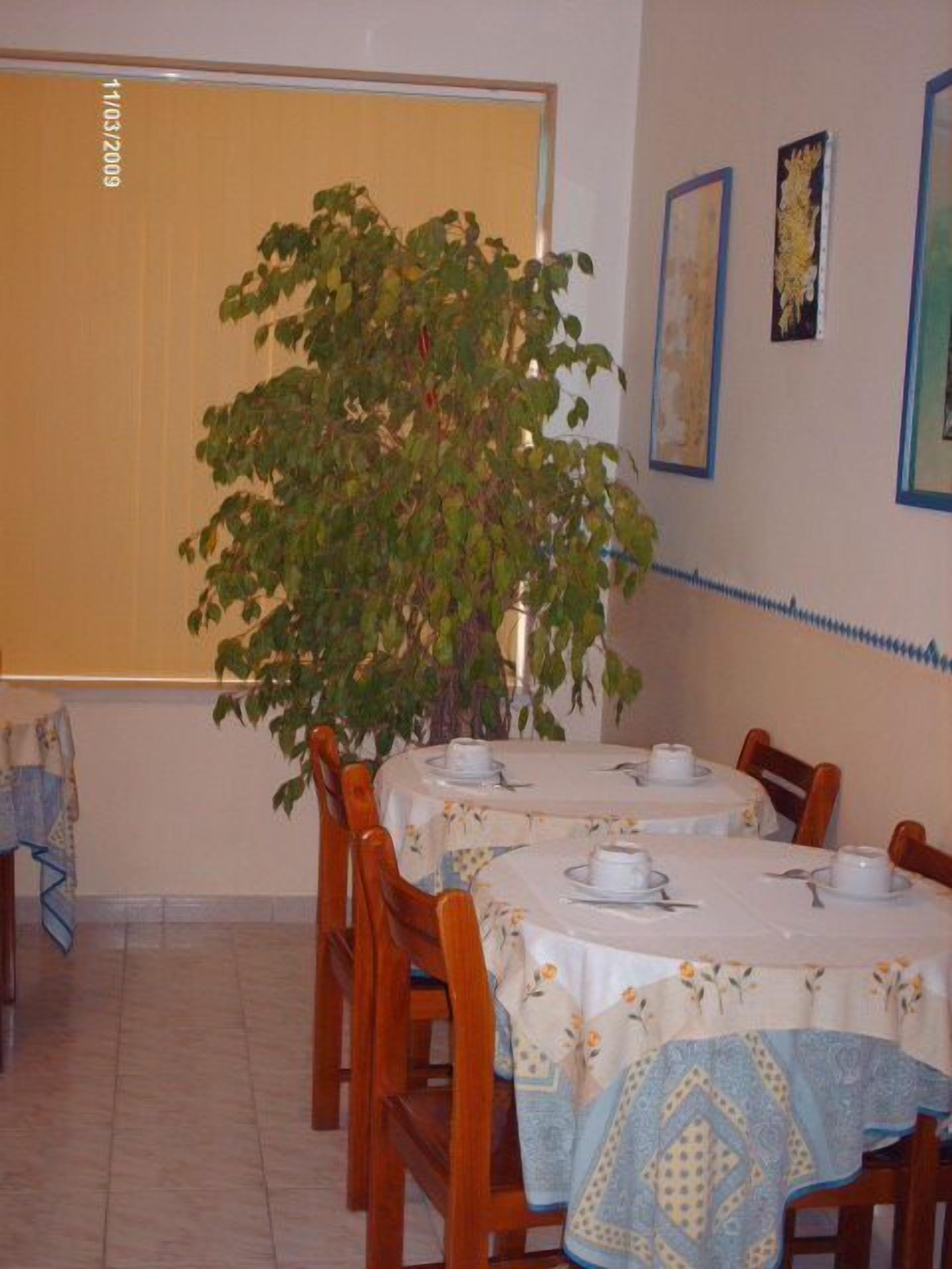 Uma sala de jantar com uma mesa posta para quatro pessoas, rodeada por cadeiras de madeira. Um vaso grande com planta está no canto, e as paredes são decoradas com obras de arte emolduradas.