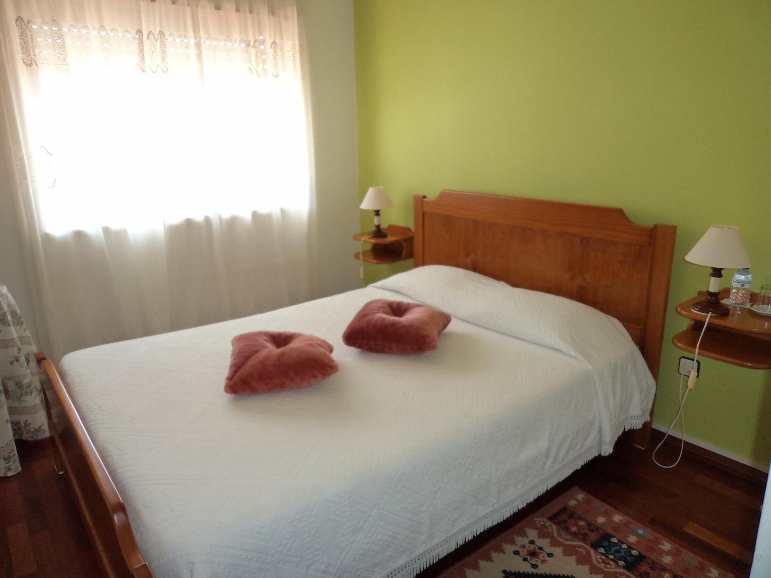 Una cama bien hecha con dos almohadas rosas, un cabecero de madera, una mesita de noche con una lámpara y una ventana con cortinas traslúcidas.