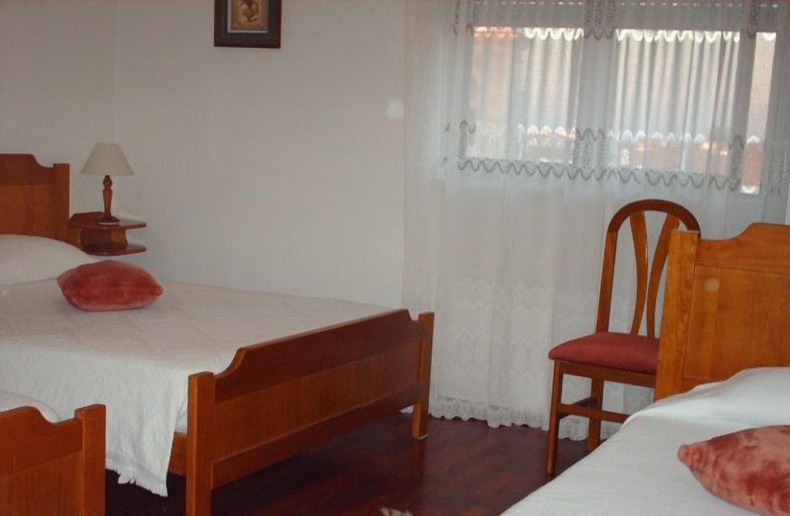 Quarto com cama de madeira, uma cadeira, mesa de cabeceira com abajur e janela com cortinas leves.