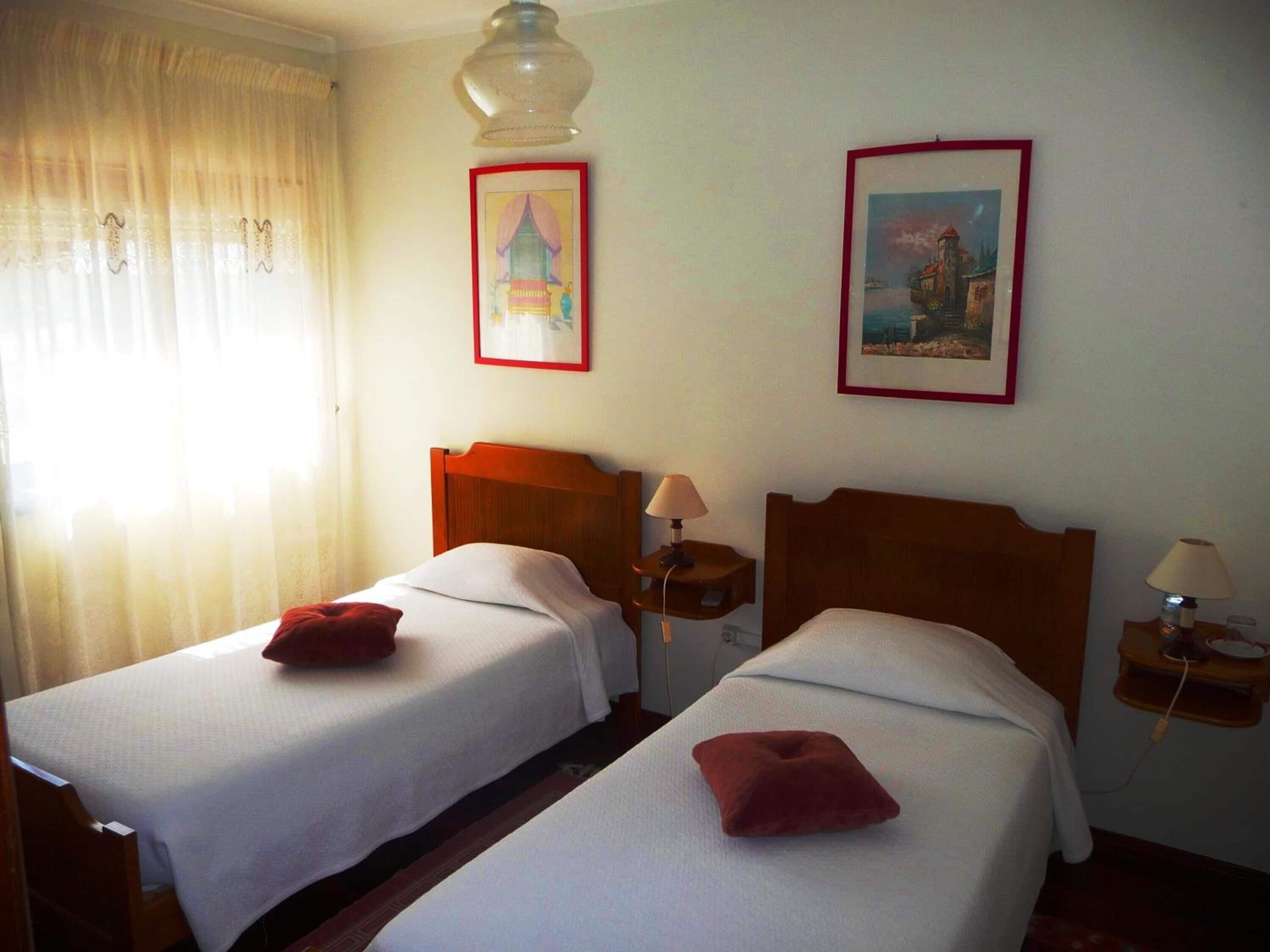 A hotel room with two single beds, each with a red pillow and a nightstand with a lamp. There are two framed pictures on the wall, one above each bed.