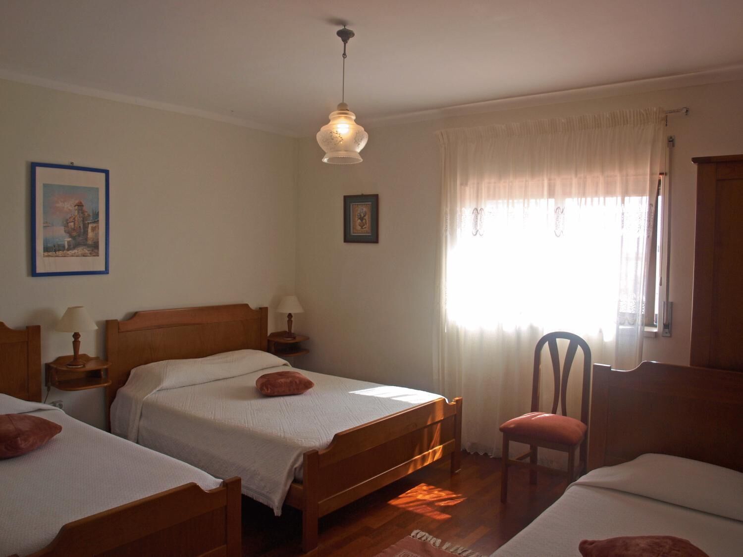 A room with two wooden beds, a wooden chair, a table with a lamp, and a hanging light fixture.