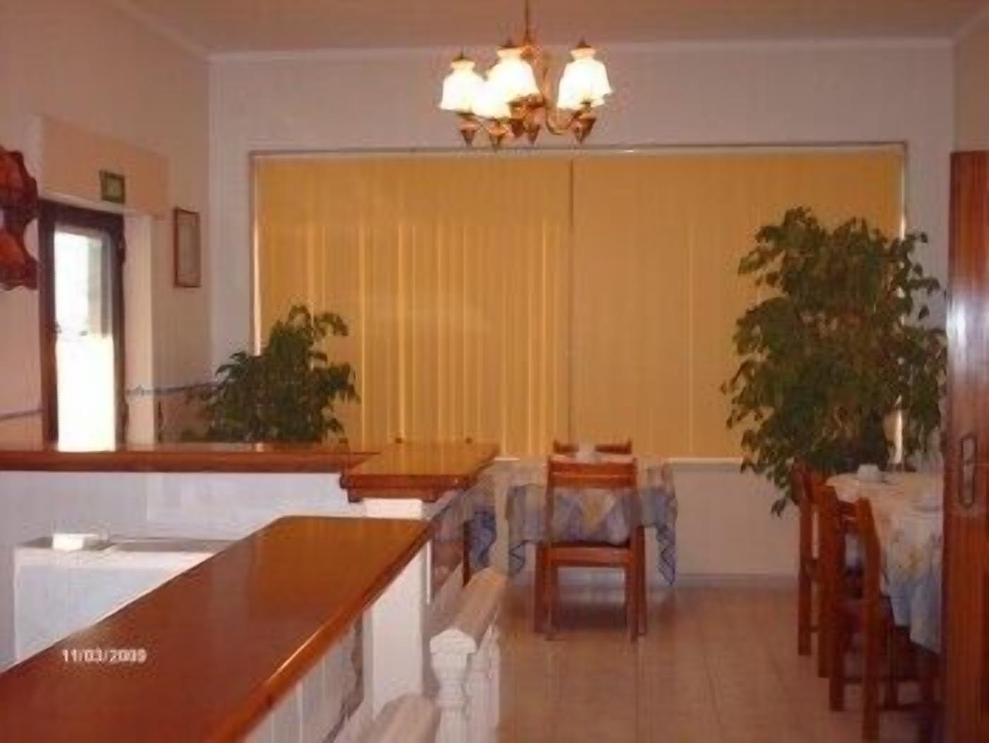 A dining area with a wooden table, chairs, and a chandelier. There are potted plants and a window with blinds.