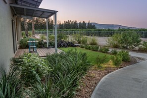 Ein überdachter Patio-Bereich mit Tisch und Stühlen, umgeben von einem angelegten Garten mit verschiedenen Pflanzen und Blick auf einen Weinberg im Hintergrund.