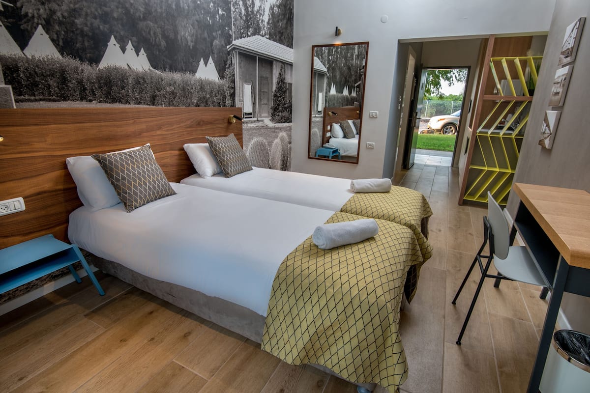 Ein Hotelzimmer mit zwei Betten, einem Holzkopfende, einem Spiegel, einem Schreibtisch und Blick nach draußen.