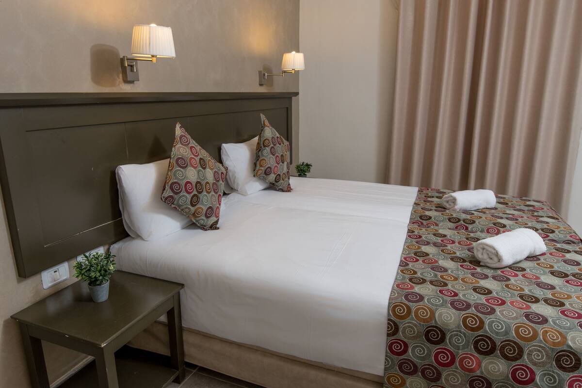 Ein Hotelzimmer mit einem großen Bett, zwei Wandleuchten, einem kleinen Nachttisch mit einer Pflanze und einem Fenster mit Vorhängen.