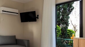 Una sala de estar moderna con un sofá gris, un televisor montado y un ventanal corredero que da a un área exterior con una terraza de madera y sillas verdes.