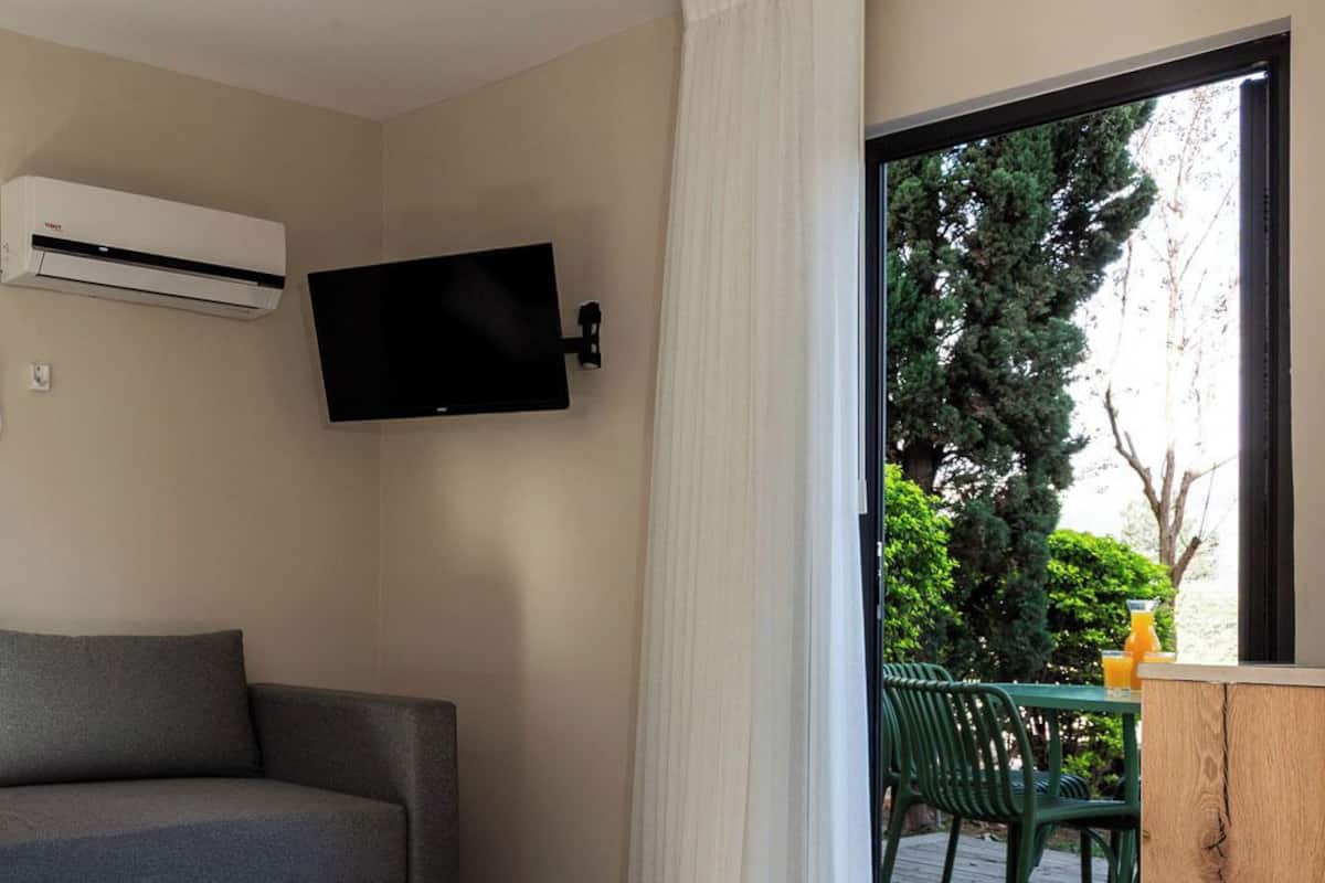 Ein modernes Wohnzimmer mit einem grauen Sofa, einem an der Wand befestigten Fernseher und einer Schiebeglastür, die zu einem Außenbereich mit Holzterrasse und grünen Stühlen führt.