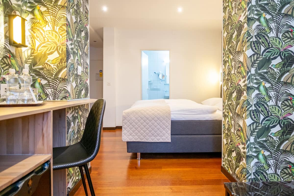 A hotel room with a bed, a desk, and a chair. There are tropical wallpaper designs on the walls.