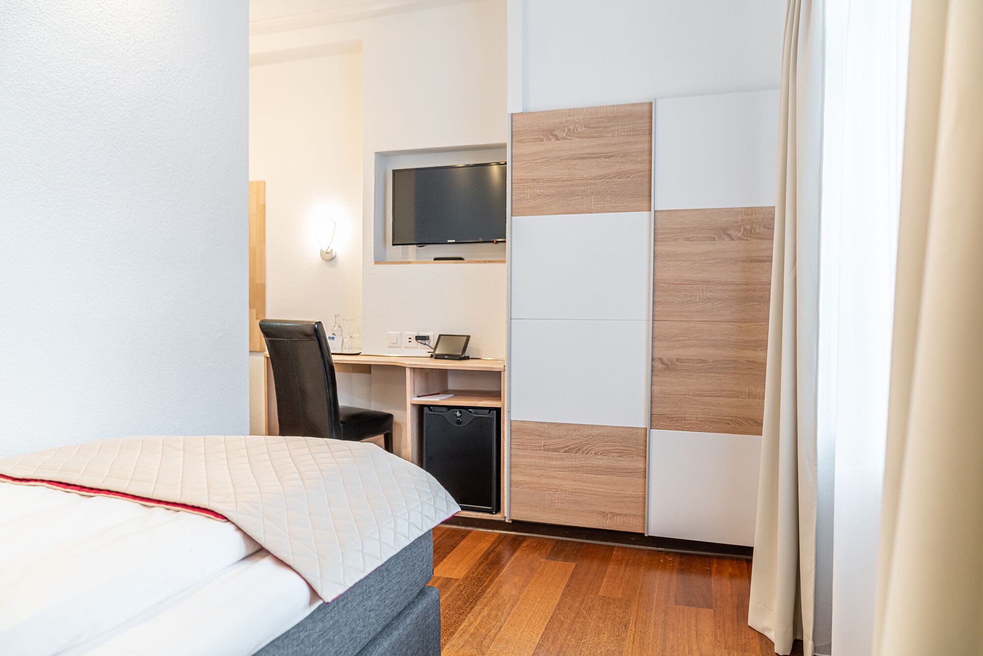 A hotel room with a bed, desk, chair, TV, and a wardrobe.