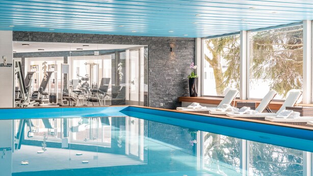 A modern indoor swimming pool with clear glass walls, a stone wall, and a view of the outdoors. - Hotel Europe Davos (Davos)