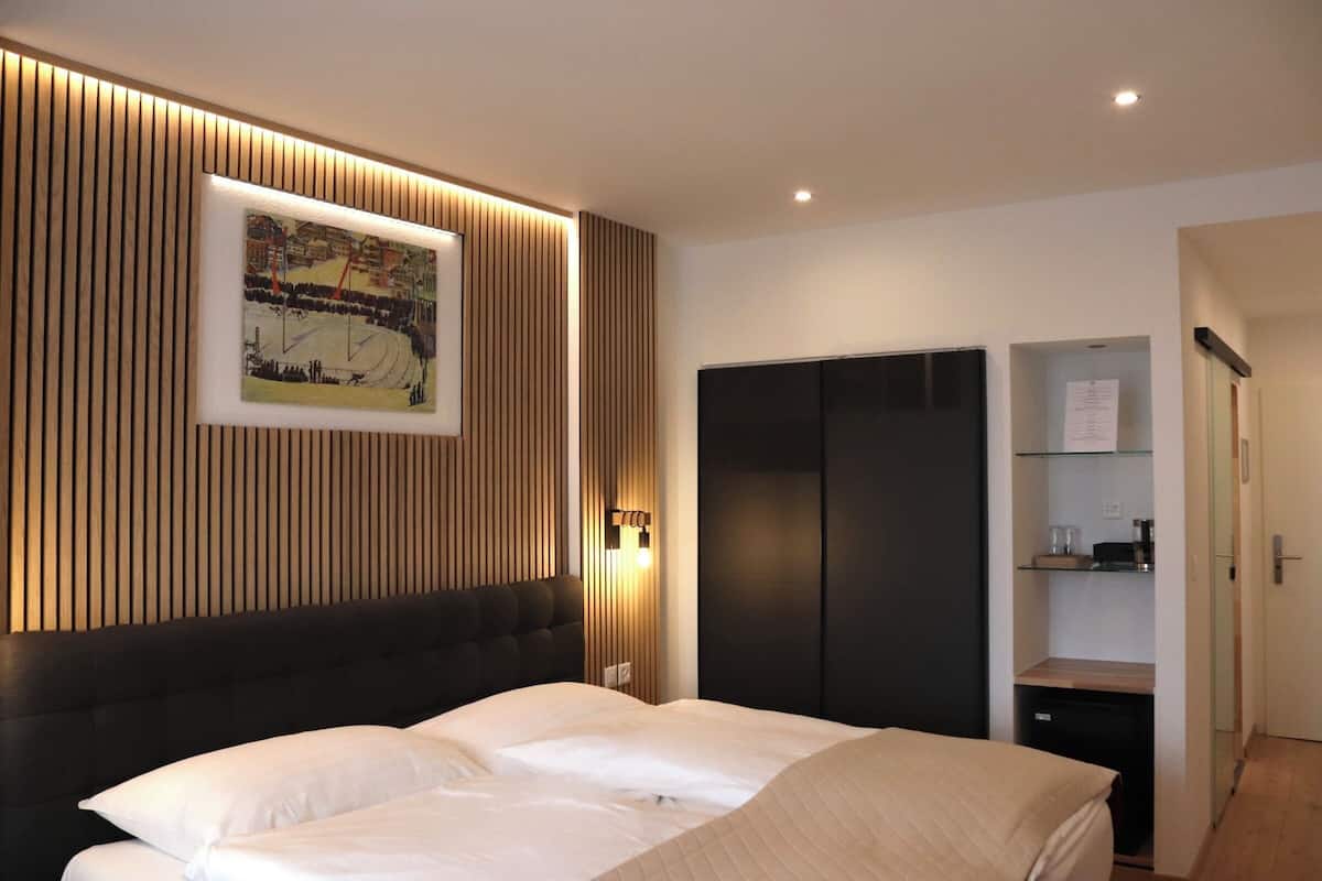 A modern hotel room with a large bed, a wooden panel wall, a framed picture, a wardrobe, and a small desk with a lamp.