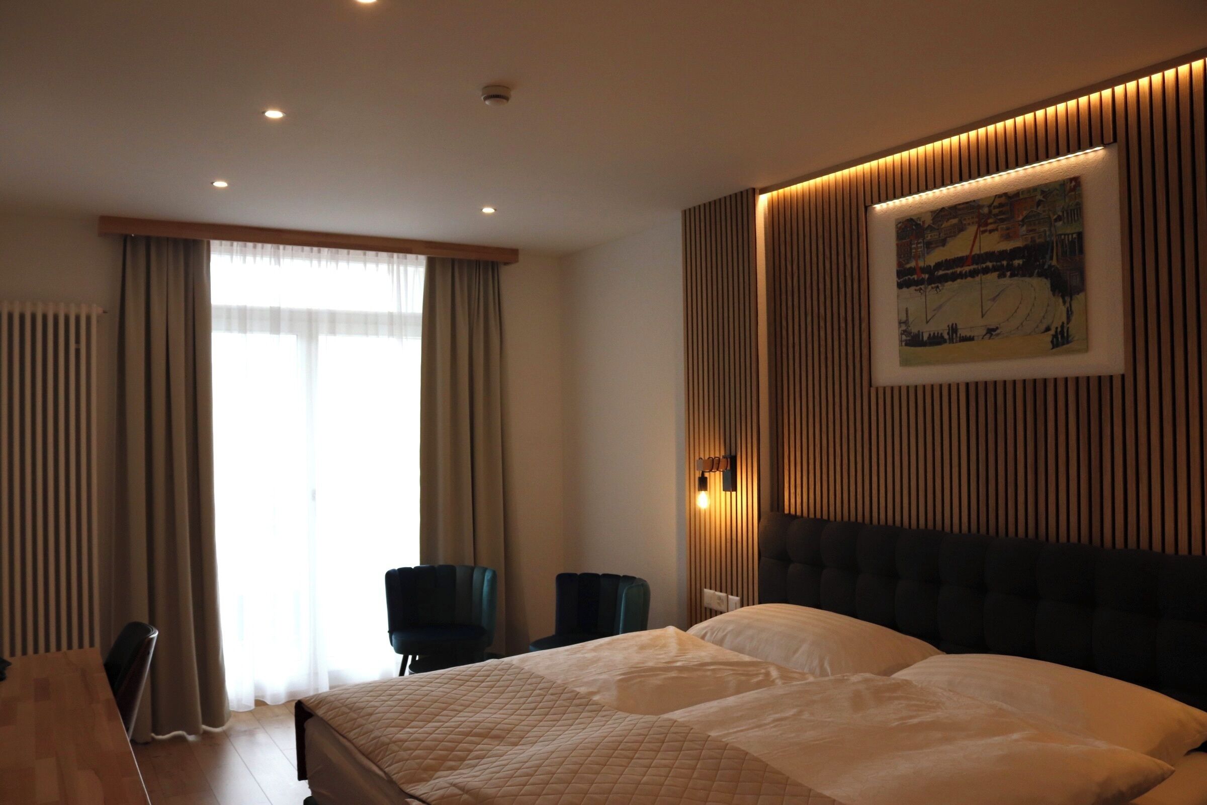 Habitación de hotel con una cama grande, un escritorio y una silla. Se ve una ventana con cortinas, una lámpara de pared y un cuadro enmarcado.
