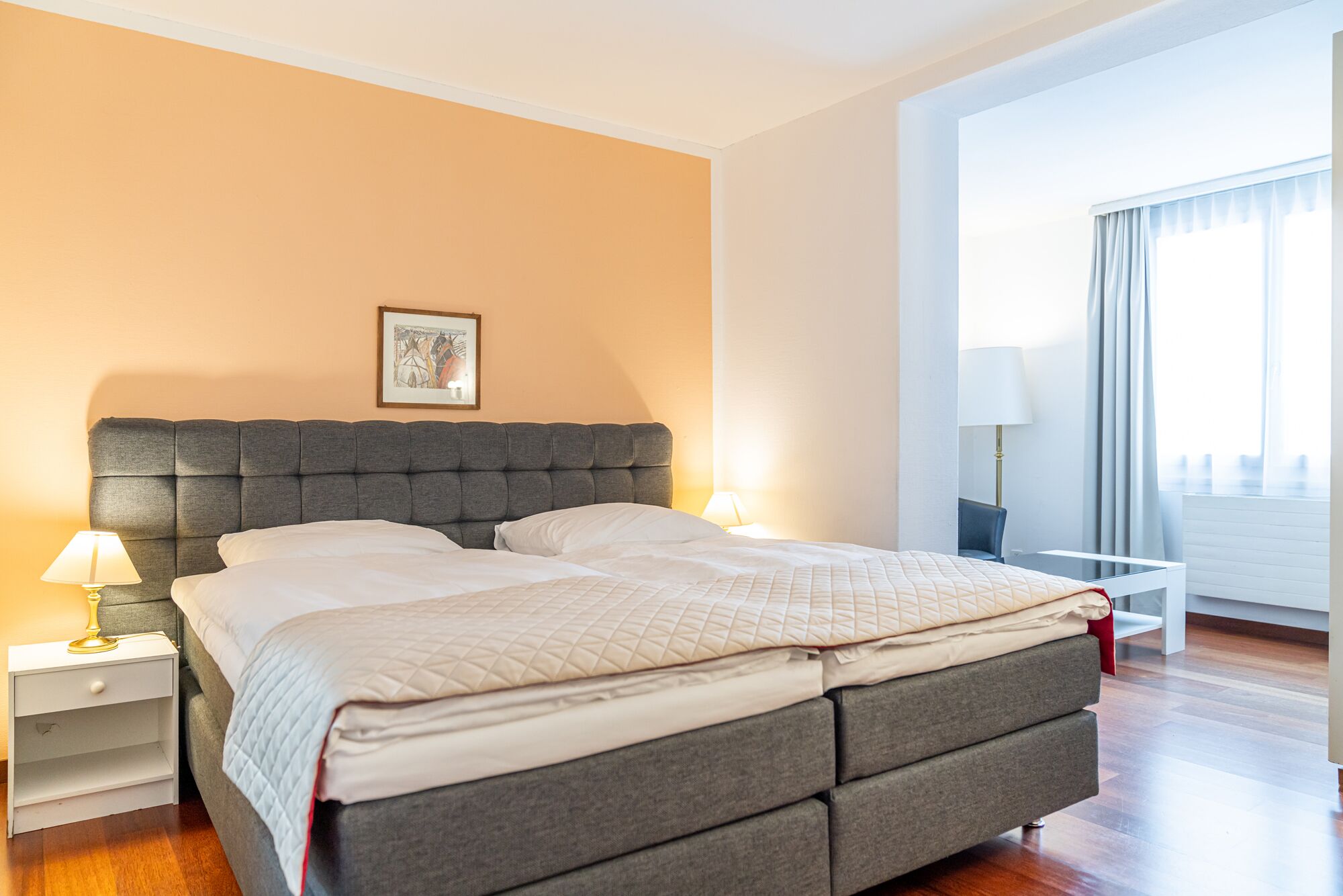 A hotel room with a large bed, a bedside table with a lamp, a small table with a lamp, and a window with curtains.