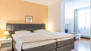 A hotel room with a large bed, a bedside table with a lamp, a small table with a lamp, and a window with curtains.