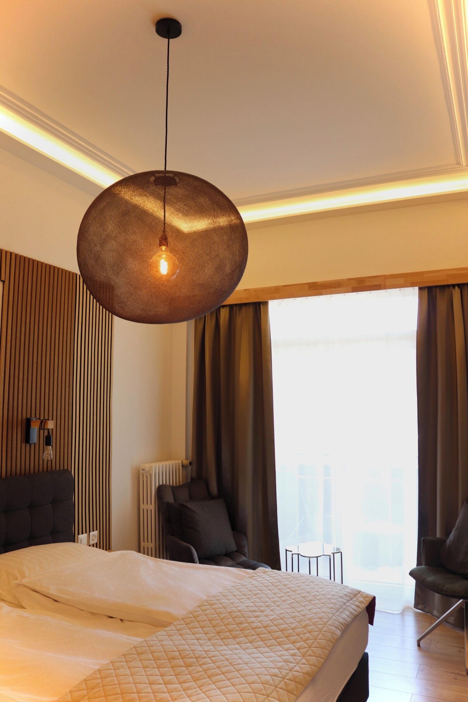 Una habitación de hotel moderna con una cama grande, una lámpara colgante, vistas de la ciudad a través de la ventana y un área de descanso con una silla y una mesa.