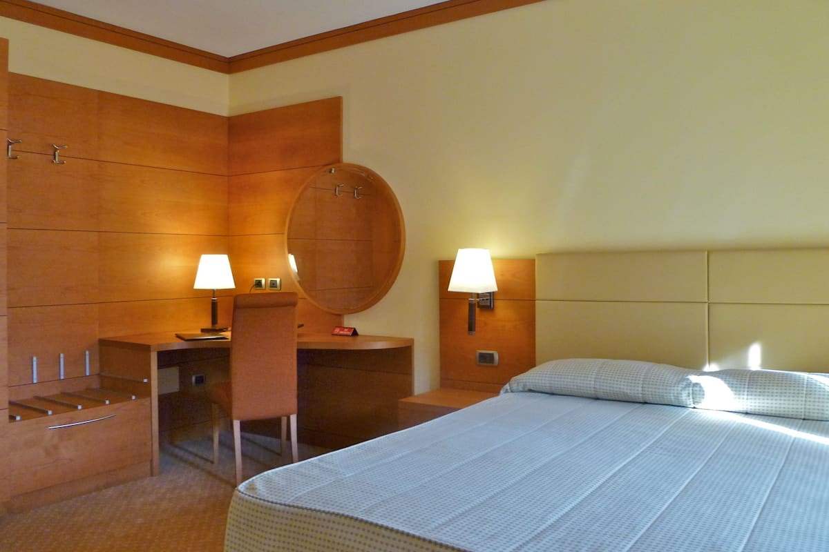 A hotel room with a bed, a desk, a chair, a round mirror, and two wall-mounted lamps.