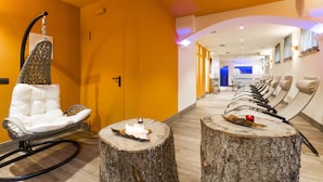 A hotel lobby with a white cushioned chair, a wooden table with a candle, and a row of sun loungers. - Relais San Giusto (Campitello di Fassa)