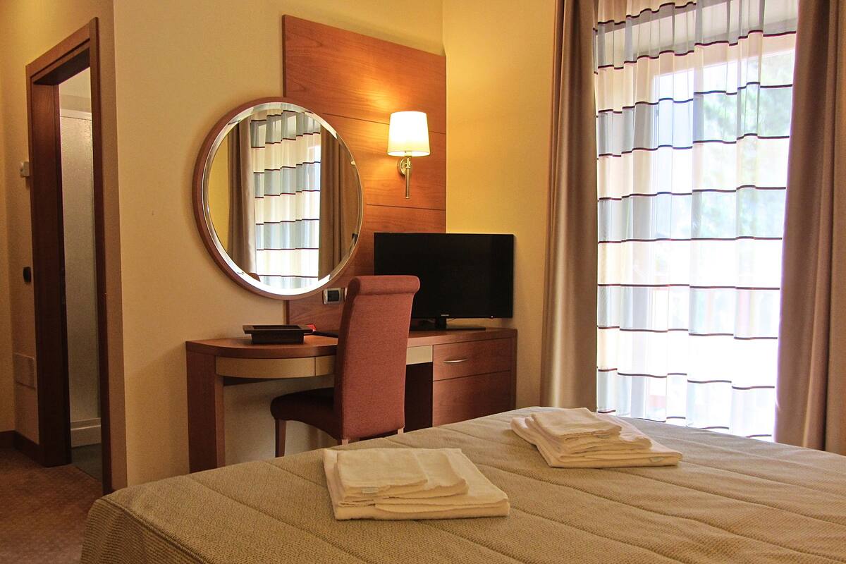 A hotel room with a bed, a desk with a chair, a television, a mirror, and a window with curtains.
