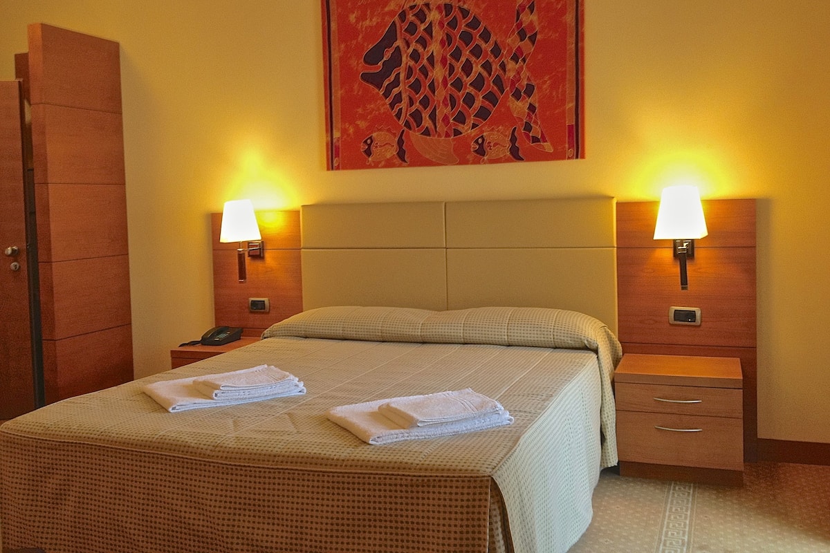 A hotel room with a bed, two bedside tables, a nightstand, a wall-mounted lamp, and a painting.