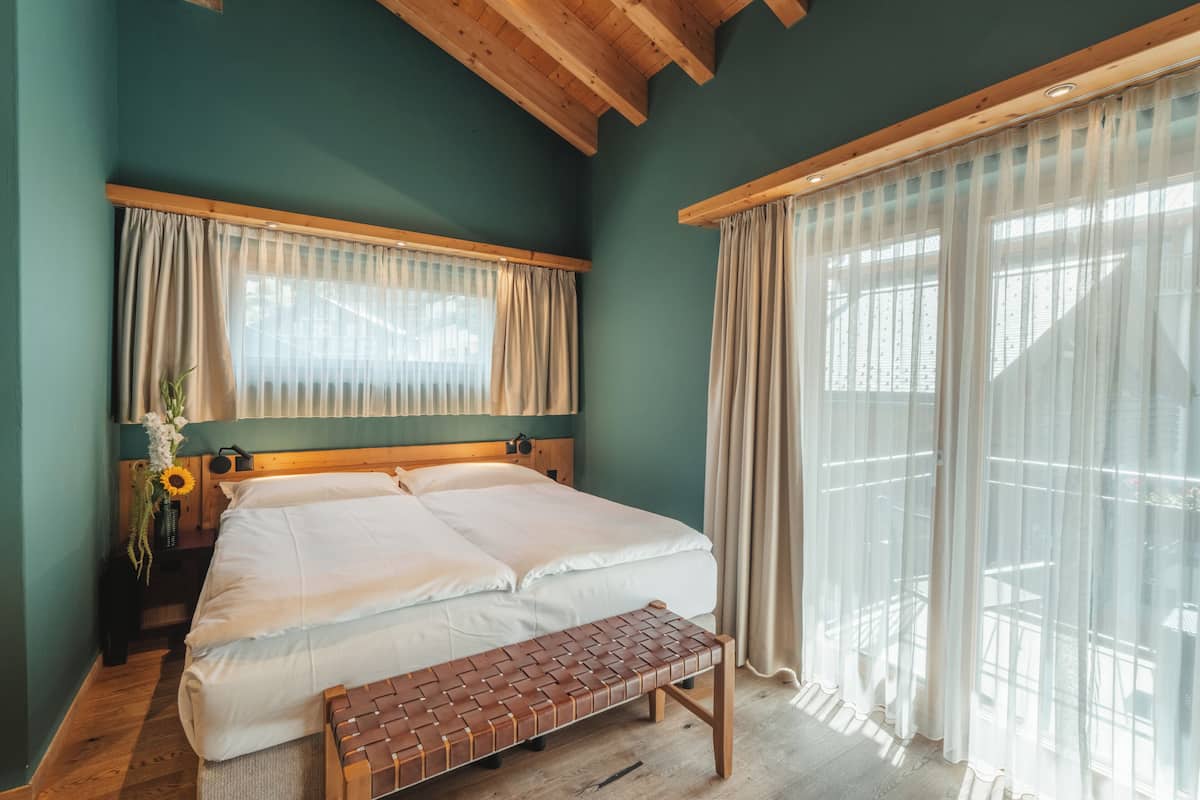 A bedroom with a bed, wooden headboard, bench, curtains, and a window.