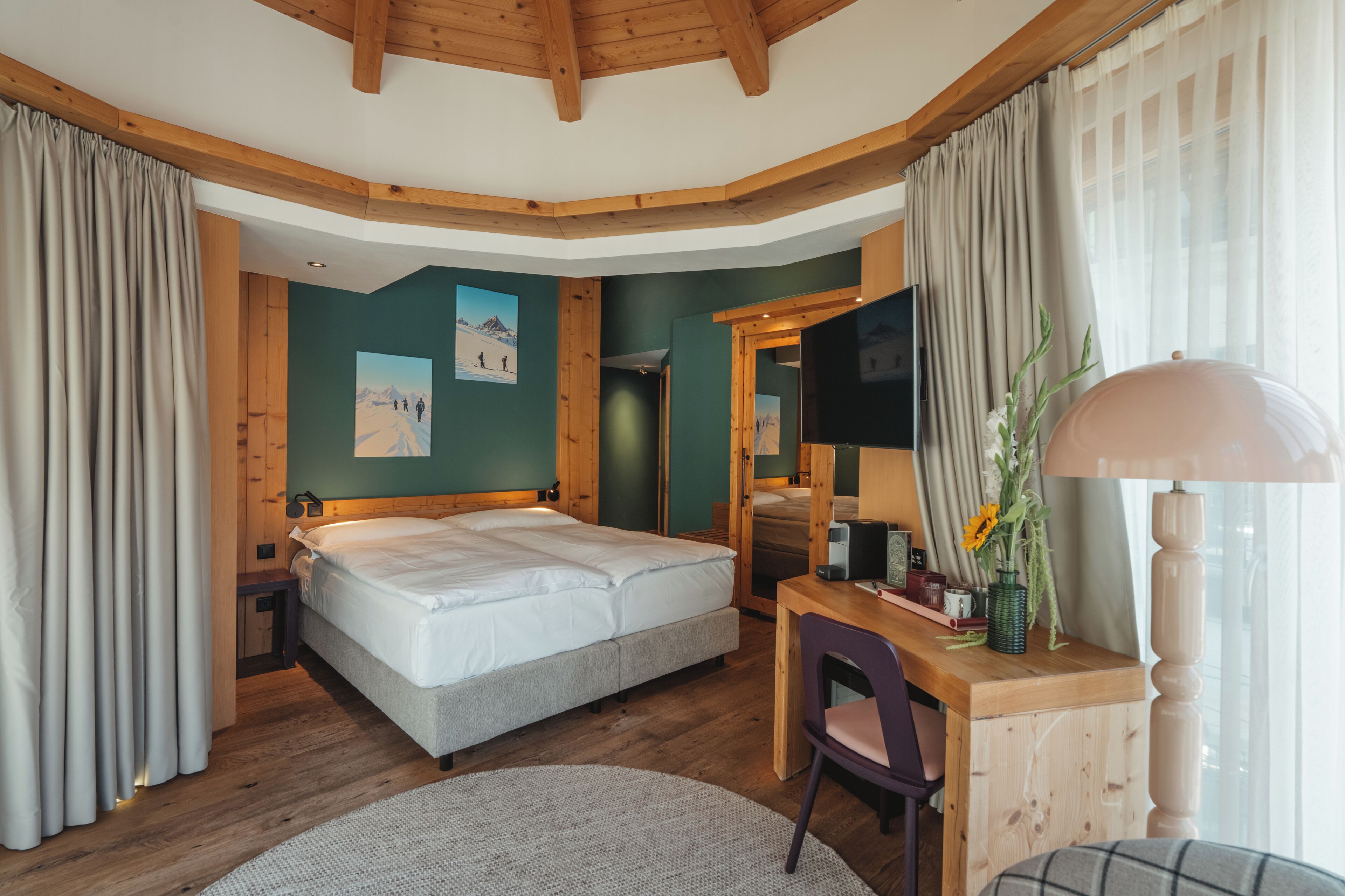 A bedroom with a bed, a desk, a chair, a TV, and a window with curtains.
