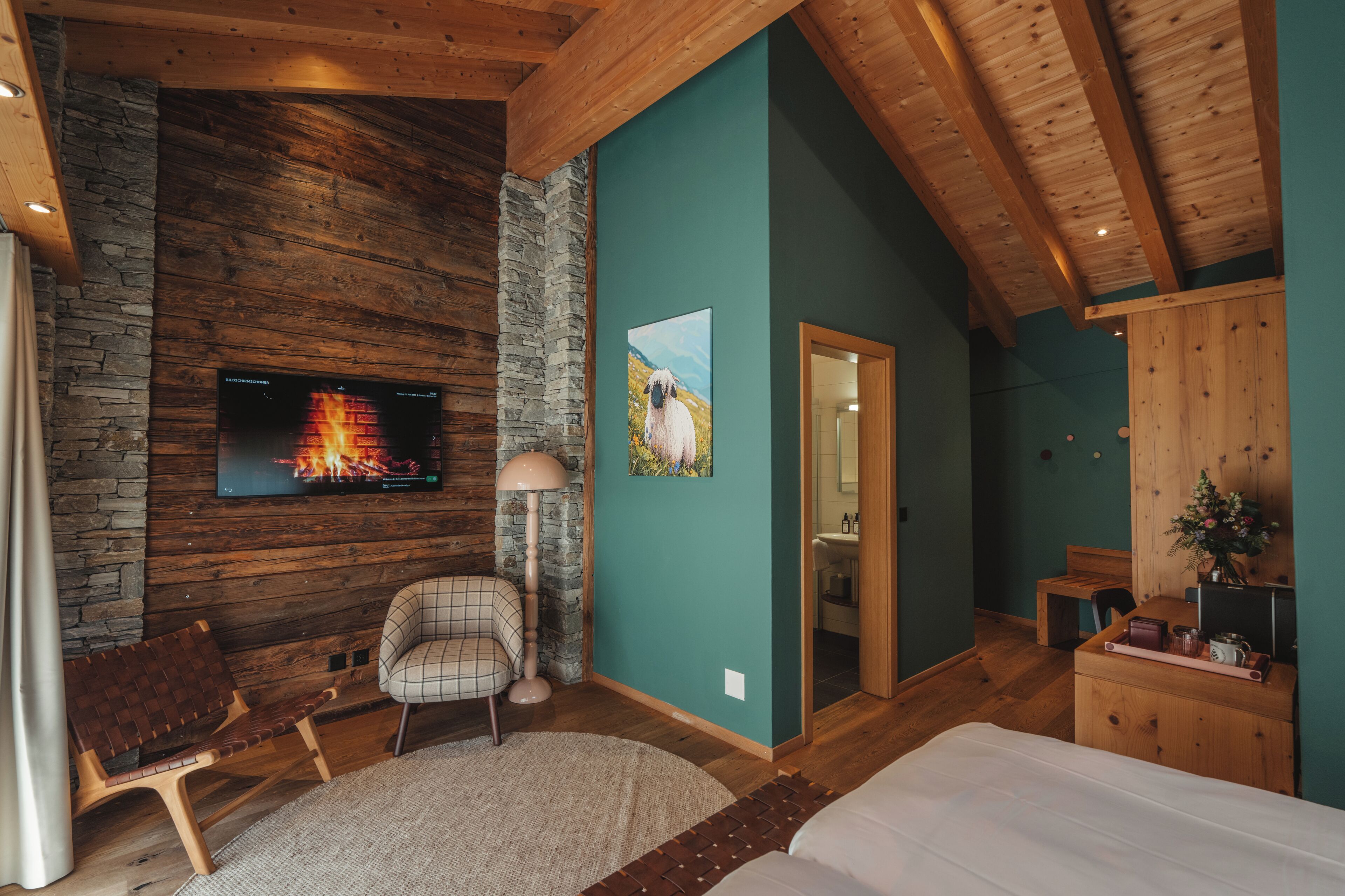 A cozy room with a stone wall, wooden ceiling, a TV mounted on the wall, a bed, a chair, and a desk with a vase of flowers.