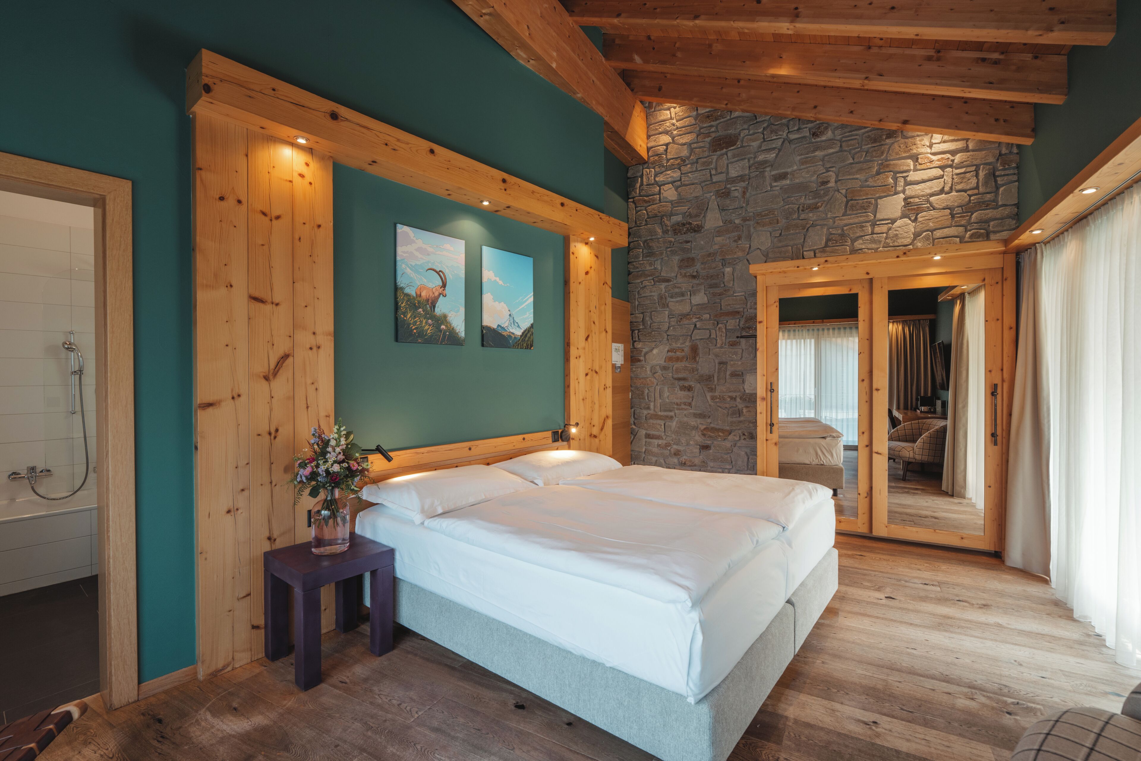 Un dormitorio con pared de piedra, vigas de madera en el techo, una cama, una mesita de noche con un florero, y un baño con ducha.