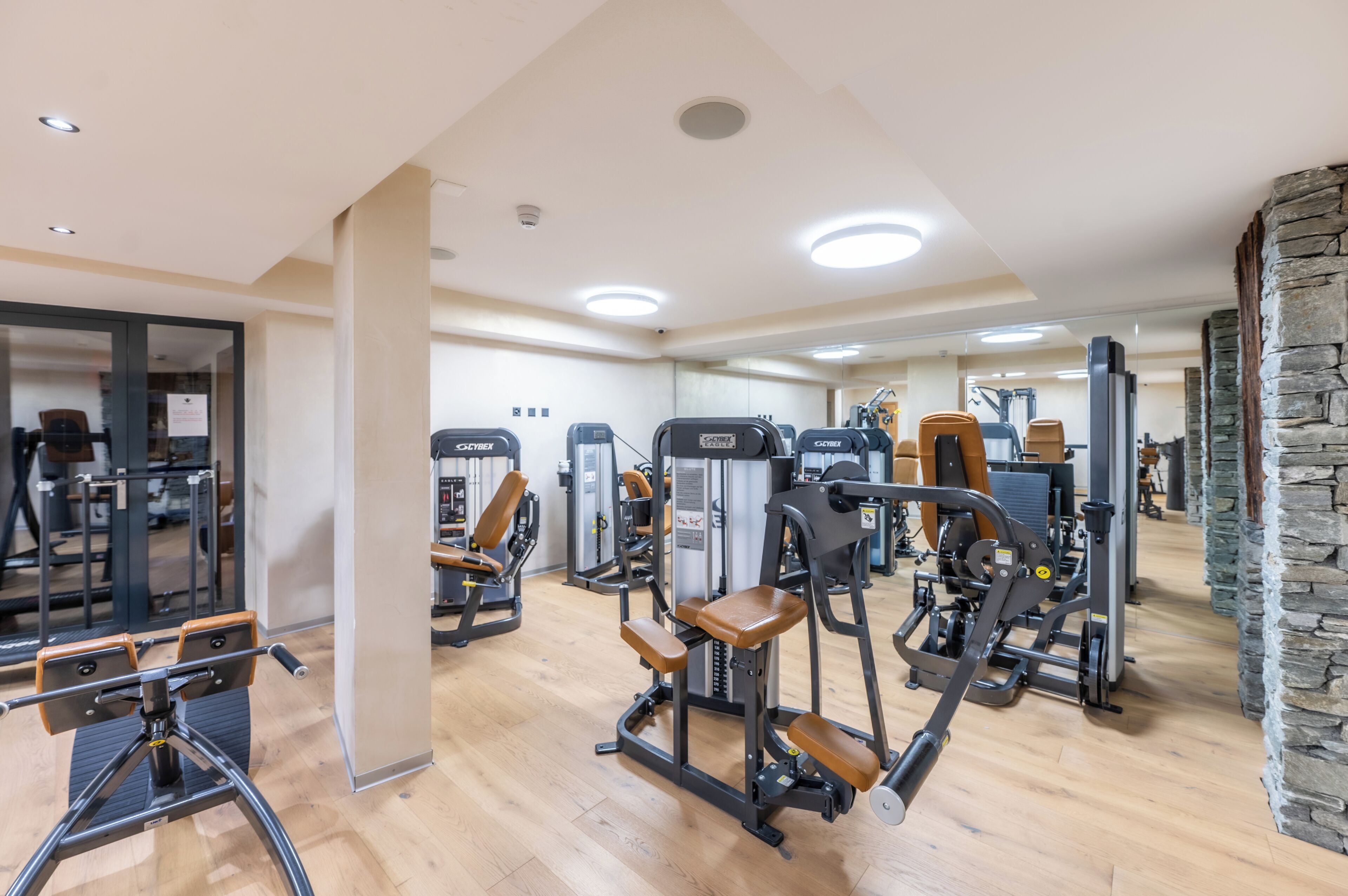 A gym with various exercise machines, wooden flooring, and a stone accent wall.