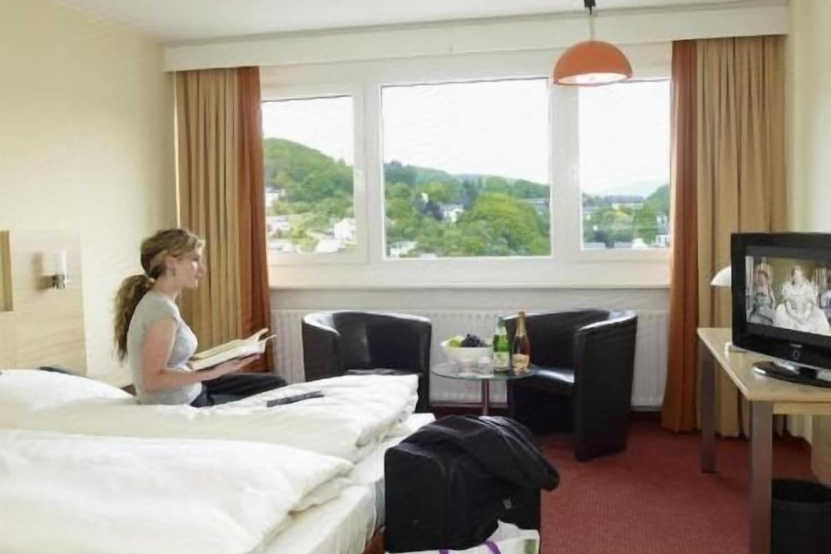 A hotel room with two beds, a television, a small table with a bottle, and a view of greenery through the window.