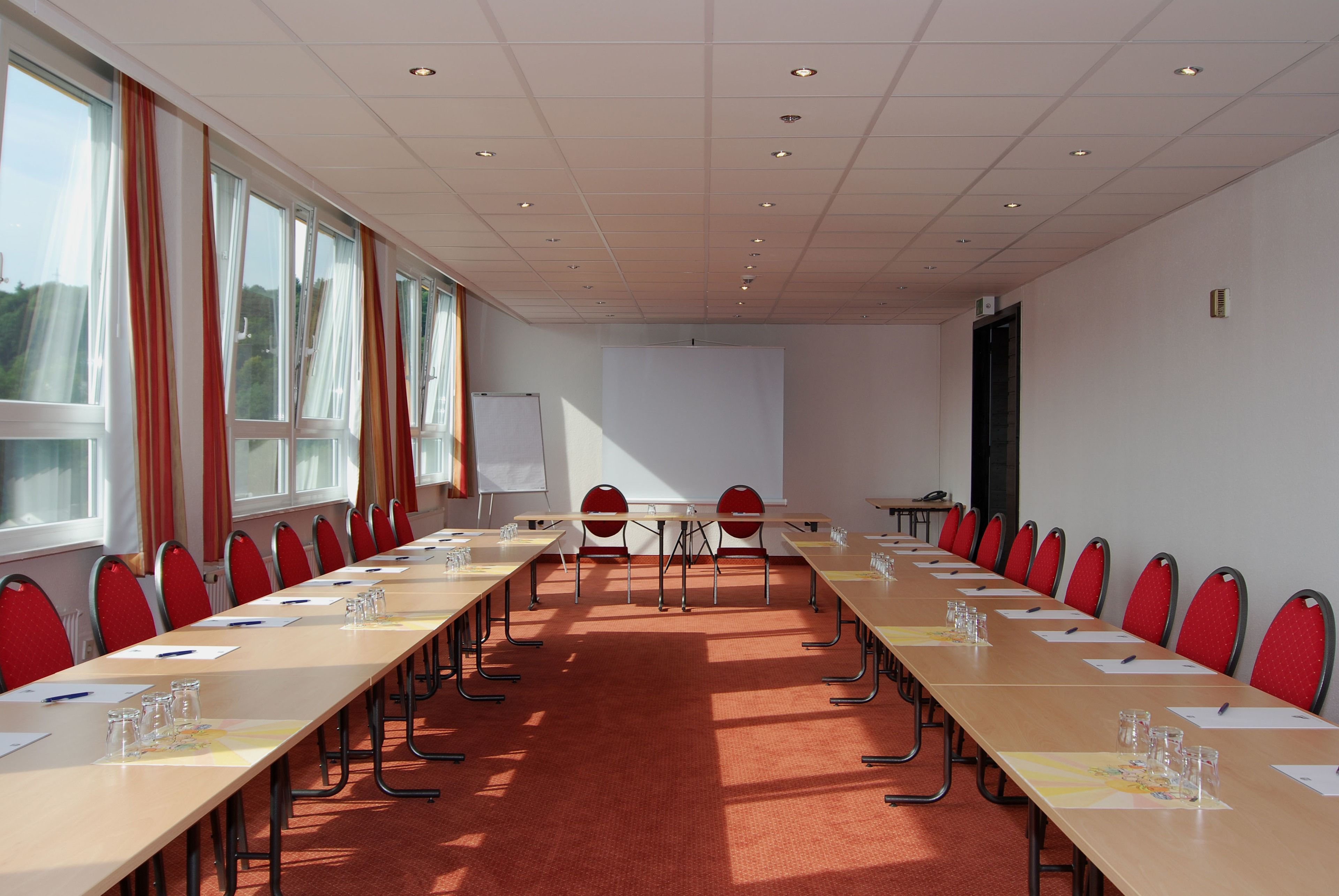 Een vergaderruimte met een lange tafel, rode stoelen en een whiteboard.