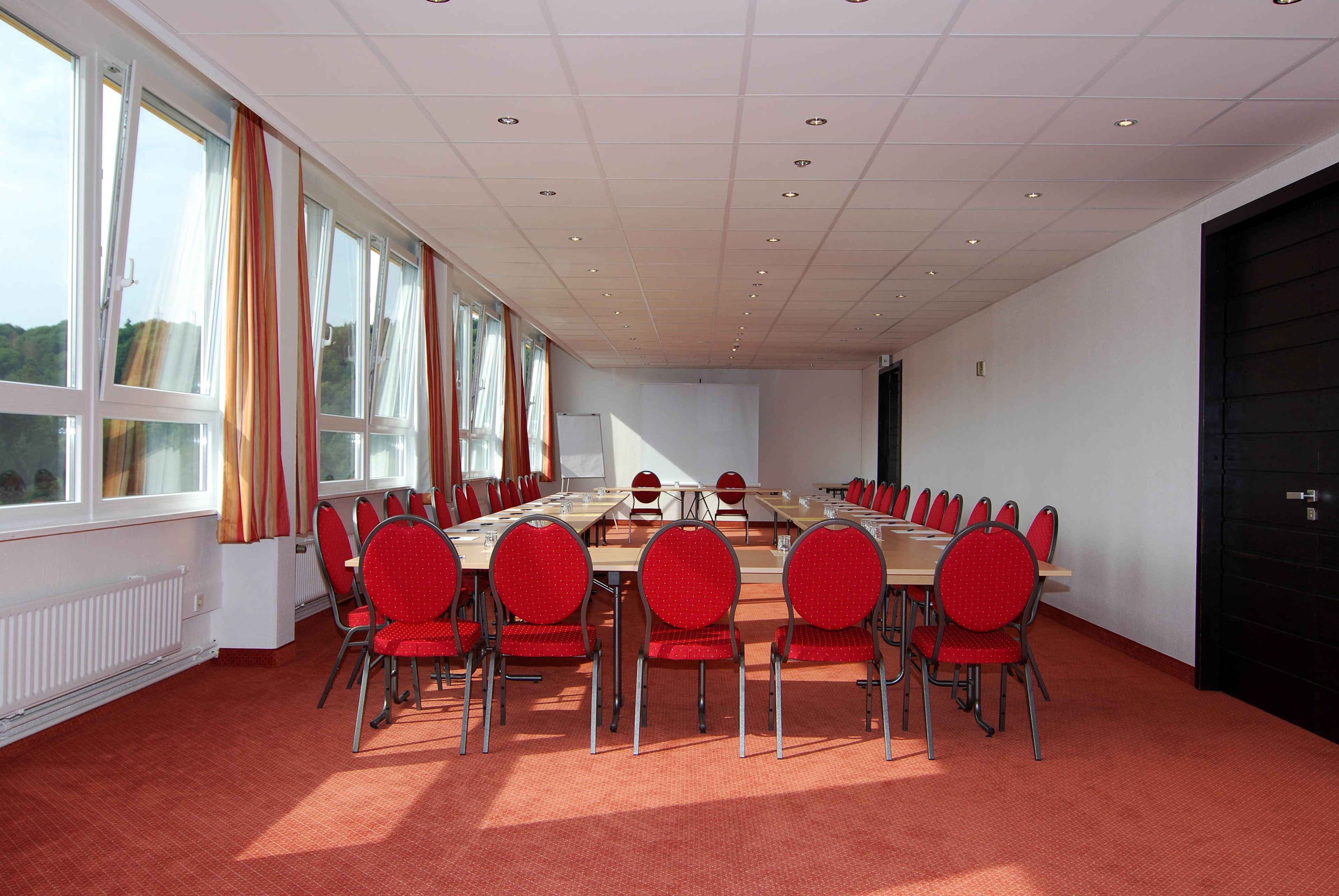 Ein Konferenzraum mit einem langen Tisch, roten Stühlen und großen Fenstern mit Vorhängen.
