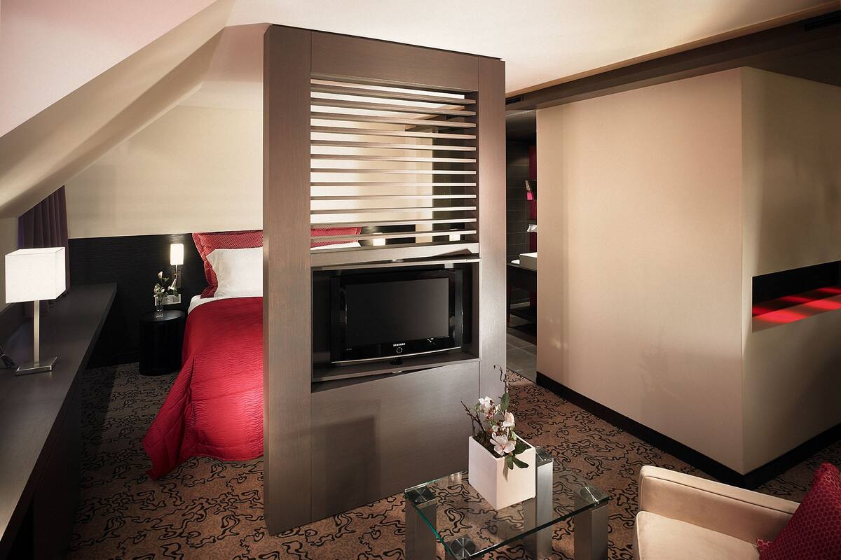A modern hotel room with a flat-screen TV mounted on a wall, a glass coffee table, a red bedspread, and a patterned carpet.