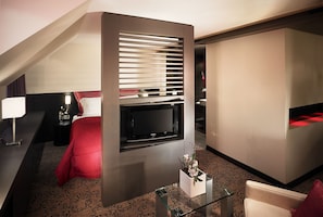 A modern hotel room with a flat-screen TV mounted on a wall, a glass coffee table, a red bedspread, and a patterned carpet.