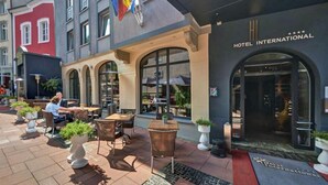 An outdoor seating area with tables and chairs, a flag, and a building with a sign reading "HOTEL INTERNATIONAL". - Hotel International (Clervaux)