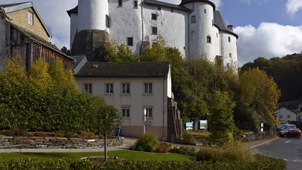 A castle-like building with multiple towers and a white facade, surrounded by greenery and a parking area. - Hotel International (Clervaux)