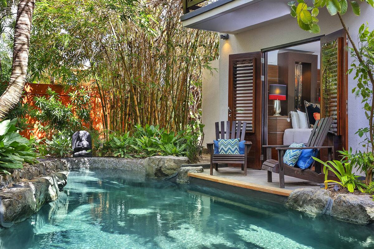 A pool area with clear water, surrounded by rocks and plants, and a wooden chair with cushions.