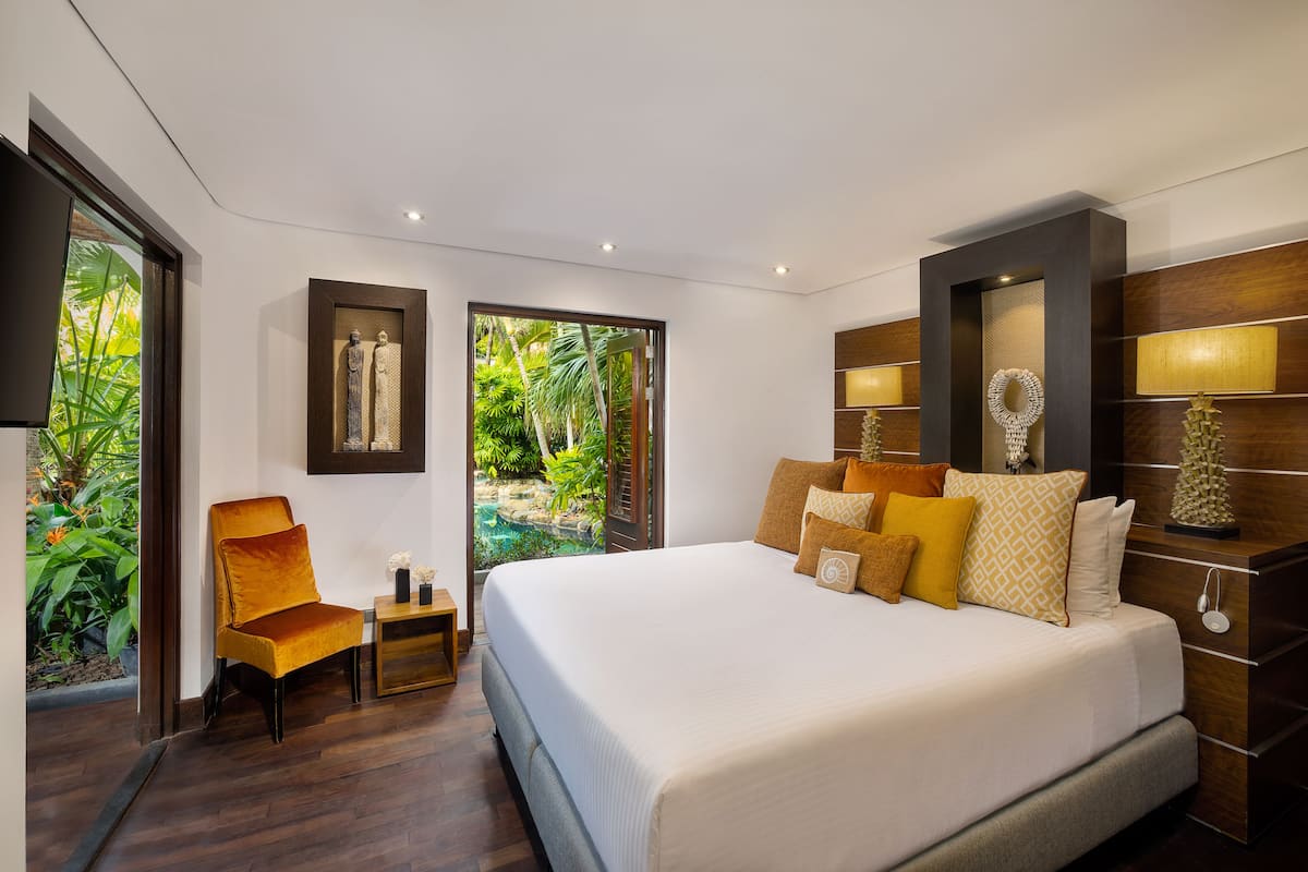 A modern bedroom with a large bed, a sitting area with a chair, and a view of a tropical garden.