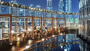 A rooftop restaurant with tables and chairs, large windows, and a city skyline view.