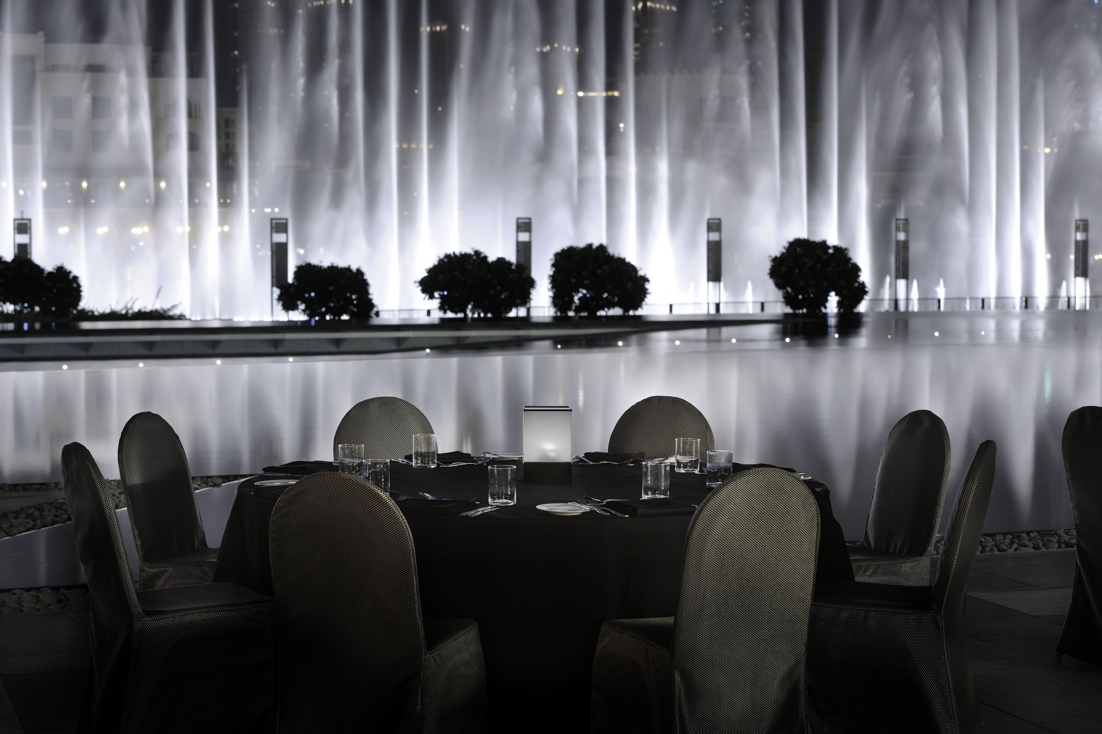 Comedor con una mesa dispuesta para cuatro personas, rodeada de sillas, con un estanque y árboles de fondo.