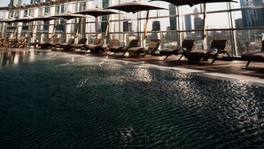 A rooftop pool area with sun loungers and umbrellas, overlooking a city skyline.
