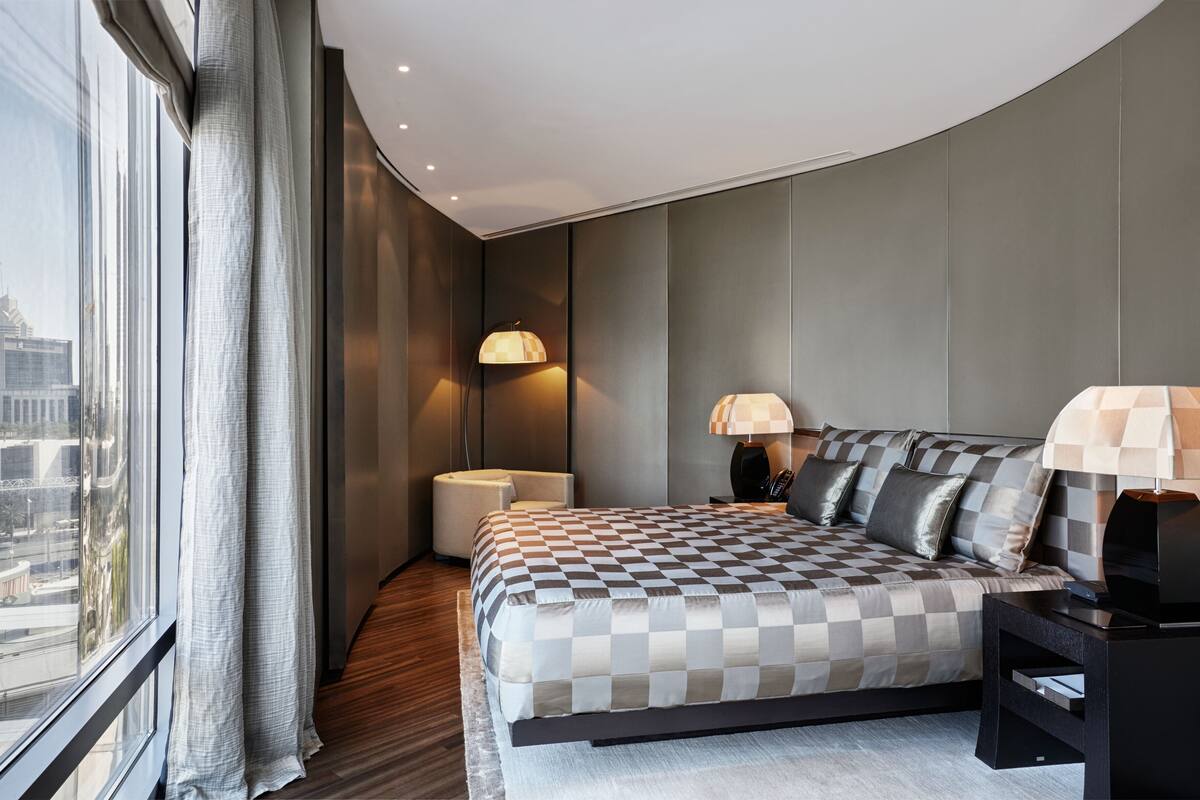 Ein modernes Hotelzimmer mit einem großen Bett, zwei Nachttischlampen, einem kleinen Nachttisch und Blick auf die Stadt durch große Fenster.